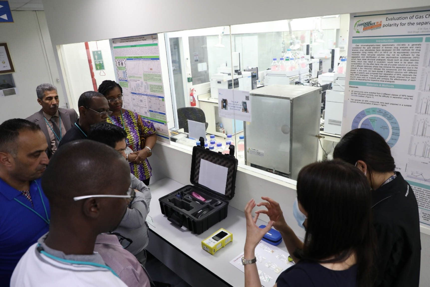 Group views scientific posters, lab equipment in open case, yellow device, and vials. Interior setting.